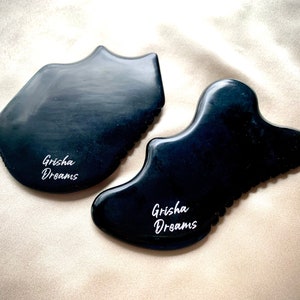 May include: Two black gua sha tools with the text "Grisha Dreams" engraved on each. The tools are made of smooth, polished stone and have a variety of curves and edges.
