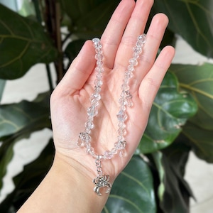 May include: A clear beaded necklace with a silver spider charm. The necklace is held in a hand, with a green leafy background. The beads are faceted and catch the light.