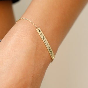 May include: Gold bar bracelet with the inscription "Type 1 Diabetes" on a woman's wrist.