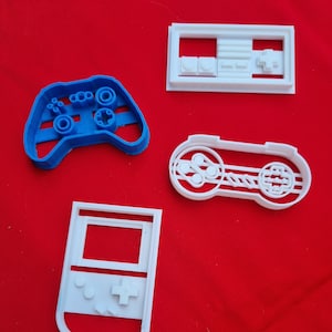 Peut inclure: Quatre emporte-pièces en plastique blanc en forme de manettes de jeu vidéo et d'une console de jeu portable. Une manette est bleue.