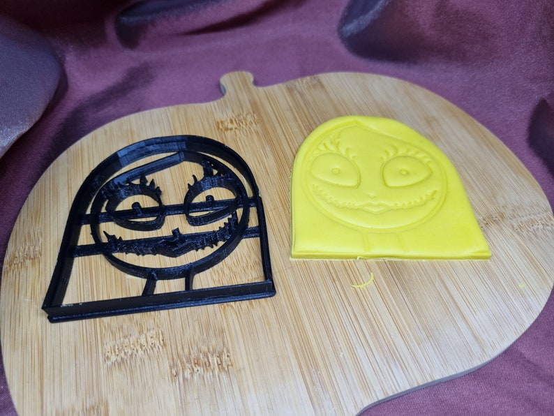 K&ouml;nnte beinhalten: Ein schwarzer Plastik-Ausstecher und ein gelber Fondant-Keks, beide in Form der Figur Sally aus dem Film Nightmare Before Christmas.