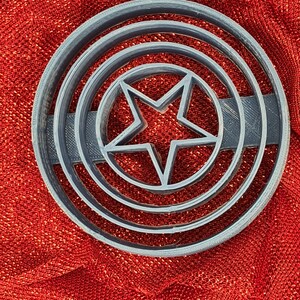 May include: A gray plastic cookie cutter shaped like a circle with a five-pointed star in the center. The circle has three concentric rings around the star.
