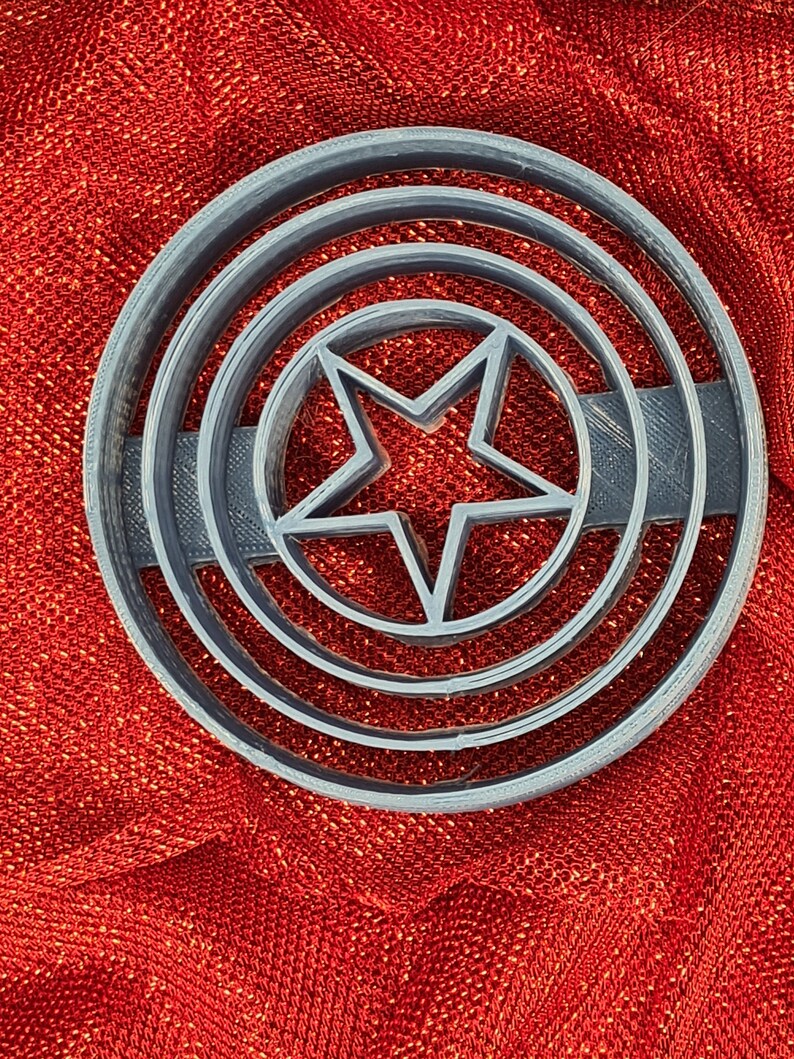 May include: A gray plastic cookie cutter shaped like a circle with a five-pointed star in the center. The circle has three concentric rings around the star.