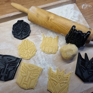 Peut inclure: Un rouleau à pâtisserie en bois et une variété d'emporte-pièces en forme de personnages Transformers, noirs et jaunes. Les emporte-pièces sont sur une surface en bois recouverte de farine blanche.
