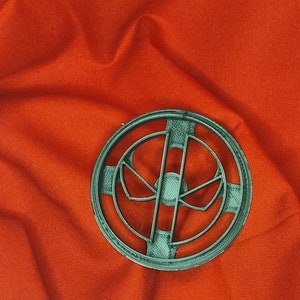 Può includere: Un emblema circolare grigio con un design stilizzato, posto su uno sfondo di tessuto rosso vibrante. L'emblema presenta un motivo geometrico all'interno di una cornice circolare.