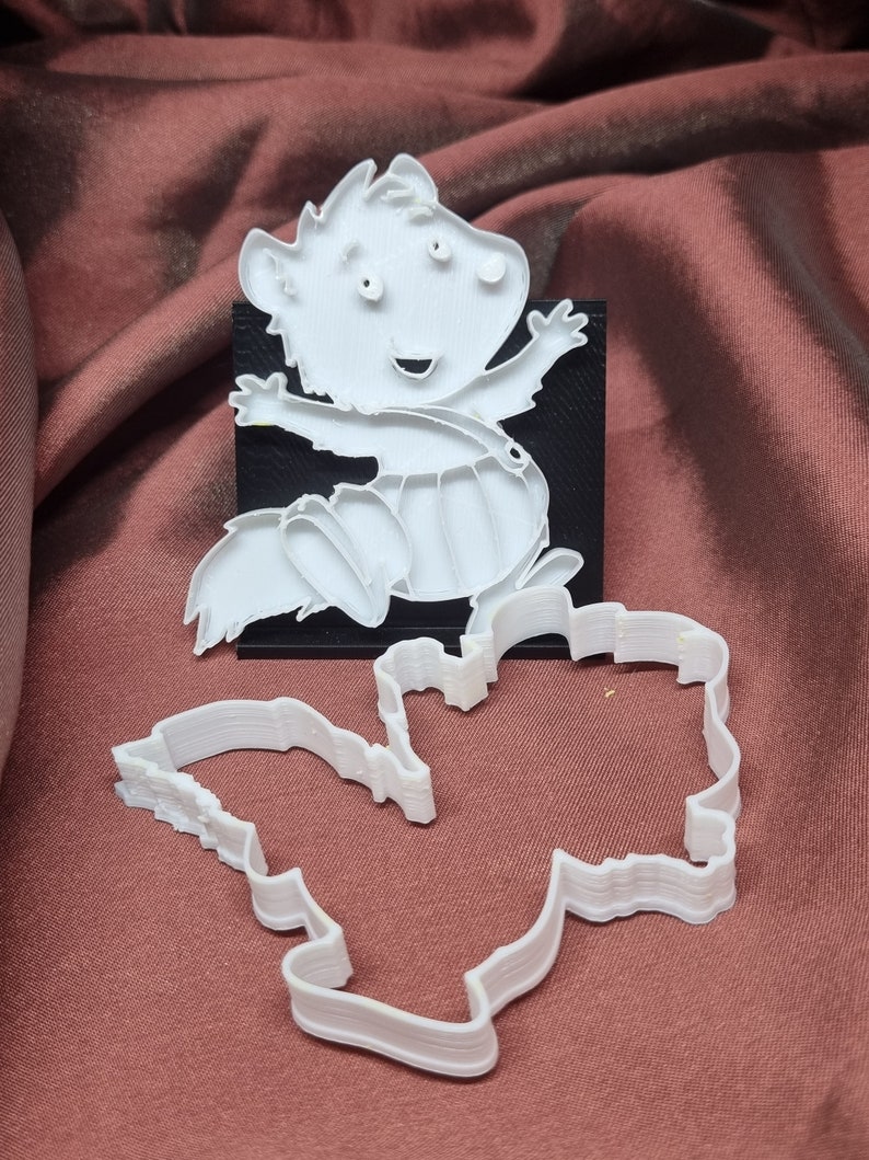 Puede incluir: Cortador de galletas de pl&aacute;stico blanco con forma de ardilla de dibujos animados. La ardilla est&aacute; de pie sobre sus patas traseras con los brazos levantados. El cortador de galletas est&aacute; sobre una superficie de tela roja.