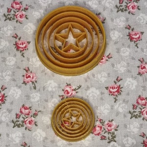 May include: Two brown plastic cookie cutters shaped like Captain America's shield. The larger cutter has three concentric circles and a five-pointed star in the center. The smaller cutter has two concentric circles and a five-pointed star in the center.