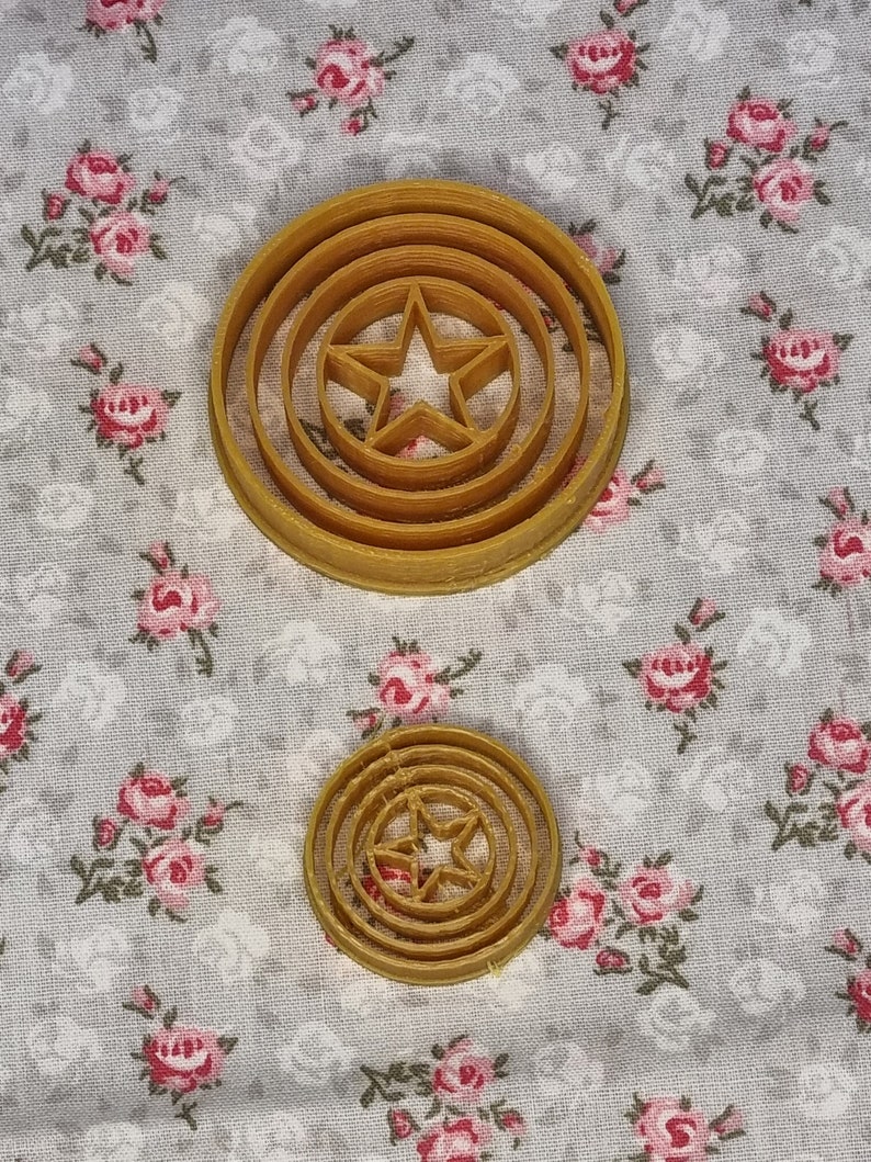 May include: Two brown plastic cookie cutters shaped like Captain America's shield. The larger cutter has three concentric circles and a five-pointed star in the center. The smaller cutter has two concentric circles and a five-pointed star in the center.