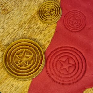 May include: Three cookie cutters in the shape of a star inside a circle. The cutters are made of yellow plastic and are on a wooden surface. Red play dough is in the background with the cookie cutters pressed into it.