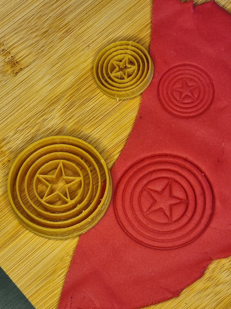 May include: Three cookie cutters in the shape of a star inside a circle. The cutters are made of yellow plastic and are on a wooden surface. Red play dough is in the background with the cookie cutters pressed into it.