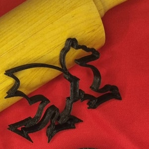 Könnte beinhalten: Schwarzer Plastik-Ausstecher in Form eines Bären. Der Ausstecher liegt auf einer roten Oberfläche neben einem Holz-Nudelholz.