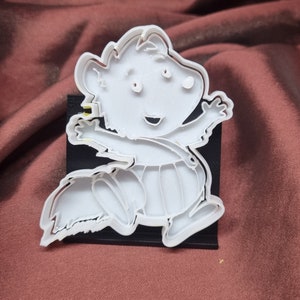Puede incluir: Cortador de galletas de pl&aacute;stico blanco con forma de personaje de dibujos animados con una gran sonrisa y ojos grandes. El personaje lleva un pa&ntilde;al y tiene una cola.