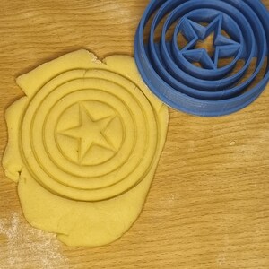 May include: A blue plastic cookie cutter shaped like a star inside a circle. The cookie cutter is on a wooden surface with a yellow dough imprint of the cookie cutter.