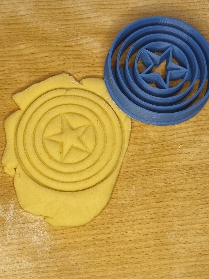 May include: A blue plastic cookie cutter shaped like a star inside a circle. The cookie cutter is on a wooden surface with a yellow dough imprint of the cookie cutter.