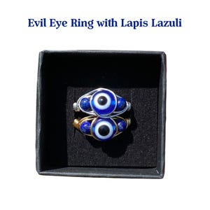 May include: Two rings with an evil eye design, set in a black box. The rings feature a blue and white eye, with blue lapis lazuli beads. One ring has a silver wire setting, and the other has a gold wire setting. Text on the image reads: Evil Eye Ring with Lapis Lazuli.