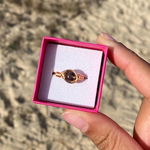 May include: A rose gold wire wrapped ring with a dark brown gemstone. The ring is displayed in a pink box.