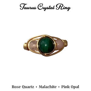 May include: A gold-toned wire ring featuring a central green malachite stone flanked by two pink stones. The ring is labeled "Taurus Crystal Ring" with the stones identified as Rose Quartz and Pink Opal.