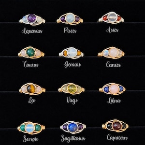 May include: Twelve wire-wrapped rings featuring various gemstones, each labeled with zodiac signs: Aquarius, Pisces, Aries, Taurus, Gemini, Cancer, Leo, Virgo, Libra, Scorpio, Sagittarius, and Capricorn. The rings use gold and silver-toned wire.