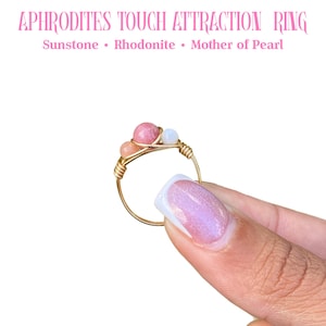 May include: A gold-colored wire ring with sunstone, rhodonite, and mother of pearl gemstones. The ring is held up against a white background. Text at the top reads "APHRODITES TOUCH ATTRACTION RING".