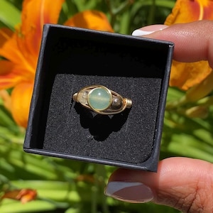 May include: A wire-wrapped ring featuring a central round light green stone, a smaller clear stone on the left, and a dark metallic bead on the right, all set within a gold-toned metal band inside a black square jewelry box.