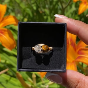 May include: A wire-wrapped ring featuring a clear, spherical crystal bead and a brown, spherical tiger's eye bead. The ring is presented inside a small, square, black box with a black velvet interior lining.