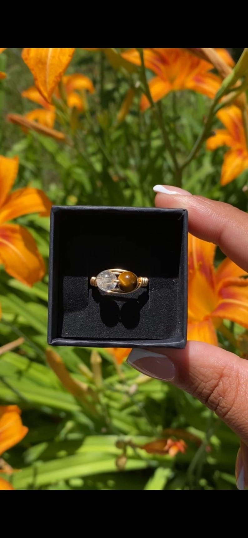 May include: A wire-wrapped ring featuring a clear, spherical crystal bead and a brown, spherical tiger's eye bead. The ring is presented inside a small, square, black box with a black velvet interior lining.