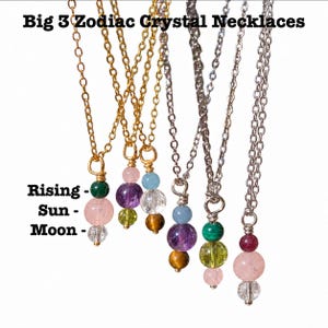 May include: Six necklaces with gold and silver chains, each featuring three stacked crystal beads. Text reads: Big 3 Zodiac Crystal Necklaces, Rising, Sun, Moon. The beads vary in color, including green, pink, clear, purple, yellow, blue, and brown.