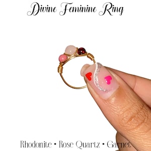 May include: A gold wire wrapped ring with pink, red, and white stones. The stones are rhodonite, rose quartz, and garnet. The ring is on a finger with red heart nail art.