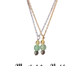 May include: Two necklaces with gold and silver chains. Each necklace features a pendant with yellow, green, and gray beads. The text "Manifestation Necklace" is below the necklaces, with the words "Citrine, Green Aventurine, Pyrite".