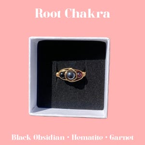 May include: A gold ring with three stones: black obsidian, hematite, and garnet. The ring is in a white box with a black interior. The text "Root Chakra" is written in white on a pink background.