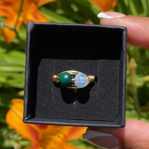 May include: A wire-wrapped ring featuring two small, round beads set in a gold-toned metal band. One bead is a solid, opaque dark green, while the other is a translucent, pale white stone. The ring is presented inside a small black box.