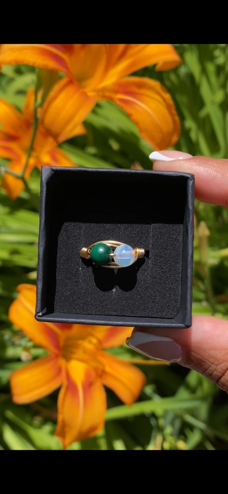 May include: A wire-wrapped ring featuring two small, round beads set in a gold-toned metal band. One bead is a solid, opaque dark green, while the other is a translucent, pale white stone. The ring is presented inside a small black box.