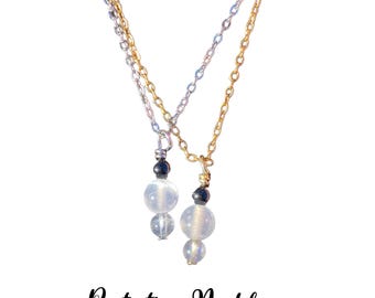 May include: Two necklaces with gold and silver chains. Each necklace has a pendant with a black bead and two clear, round stones. The text "Protection Necklace" is below the necklaces, with "Black Tourmaline • Selenite • Labradorite" underneath.