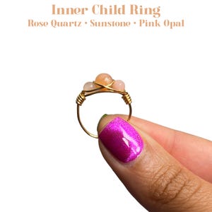 May include: Gold wire ring with three pink and orange gemstones. The ring is labeled "Inner Child Ring" and "Rose Quartz, Sunstone, Pink Opal".