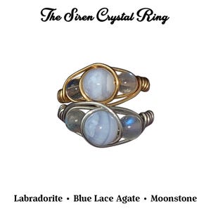 May include: Two wire-wrapped rings with a central blue lace agate stone and smaller labradorite and moonstone accents. The top ring has gold wire, the bottom ring has silver wire. Text reads "The Siren Crystal Ring".