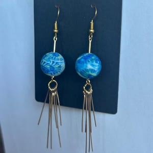 Puede incluir: Un par de pendientes colgantes con ganchos de metal dorado y flecos de hilos metálicos dorados. Cada pendiente incluye una cuenta esférica con un patrón jaspeado en tonos azul brillante, azul claro y blanco.