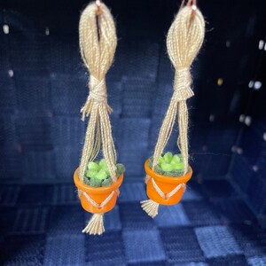 Könnte beinhalten: Ein Paar handgefertigte Ohrringe mit Miniatur-Hängepflanzen mit grünen Filz-Sukkulente und orangefarbenen Töpfen. Die Pflanzen hängen an beigefarbenem Garn.