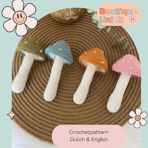 Op de afbeelding: Vier gehaakte paddenstoelspeelgoed in groen, blauw, oranje en roze met witte stelen. De paddenstoelen zijn gerangschikt op een geweven, cirkelvormige mat. De afbeelding bevat de tekst "Boutique Uniek" en "Crochetpattern Dutch & English".