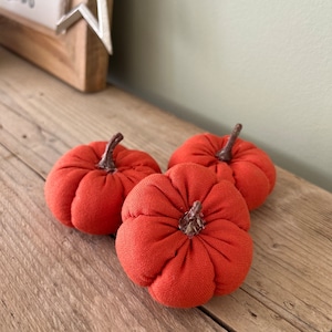 May include: Three orange fabric pumpkins with brown stems. The pumpkins are sitting on a wooden surface.