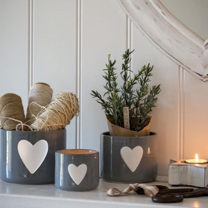 May include: Three grey ceramic planters with white heart designs. The planters are filled with twine and a small potted plant with a "Rosemary" label.