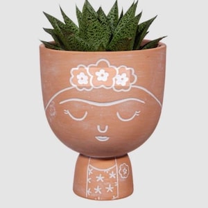 May include: A terracotta ceramic plant pot in the shape of a face, adorned with a floral crown and closed eyes. It contains a green succulent plant. White paint details the face and floral design. The pot has a pedestal base with a floral pattern.