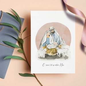 Op de afbeelding: Een aquarel-illustratie van de kerststal met Maria, Jozef en baby Jezus. De scène speelt zich af tegen een roze en witte achtergrond met witte schapen rondom de familie. De tekst "O come let us adore Him" staat onder de afbeelding.