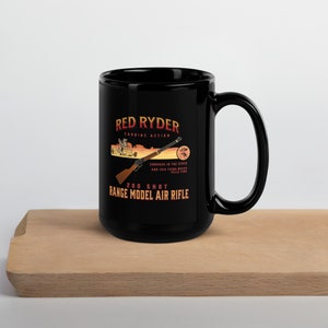May include: Black ceramic mug featuring a white graphic of a Red Ryder Carbine Action 200 Shot Range Model Air Rifle. The graphic includes a compass in the stock and the text "Compass in the stock and this thing which tells time".