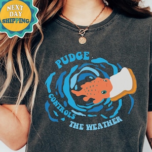 以下が含まれることがあります： パンを食べる漫画の魚のグラフィックがプリントされたダークグレーのTシャツ。魚を囲むように「Pudge Controls The Weather」というテキストがプリントされています。