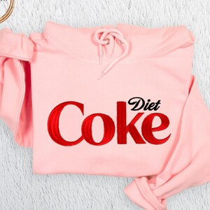 May include: A light pink hooded sweatshirt featuring the red embroidered text 'Coke' and the black embroidered text 'Diet' centered on the front. The garment includes a matching drawstring hood and long sleeves, presented with a circular logo.