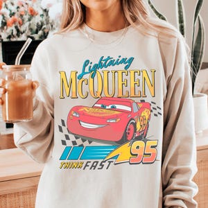 A person wearing a white sweatshirt with a graphic of Lightning McQueen from the Cars movie, holding a drink.