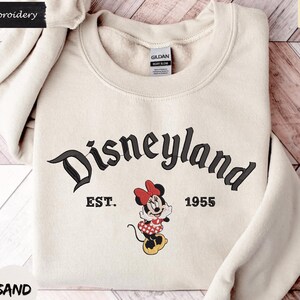 A beige sweater with the text "Disneyland EST. 1955" and a Minnie Mouse graphic printed on it, along with the text "SAND" at the bottom.
