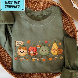Peut inclure: Un sweat-shirt à col rond vert olive orné d'un imprimé graphique représentant des produits d'automne anthropomorphes, dont une citrouille tenant un drapeau avec l'inscription HELLO FALL, une pomme avec une tasse, une citrouille verte et une poire.
