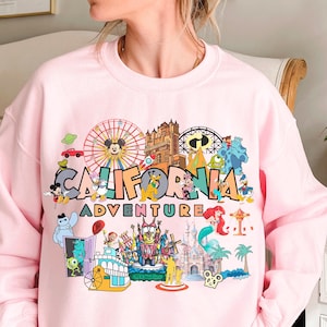A woman wearing a pink sweatshirt with a colorful graphic design depicting various Disney and amusement park attractions, including Mickey Mouse, the California Adventure, and other iconic landmarks and characters.