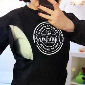 May include: Black long-sleeved shirt with a white graphic that reads "Mama's Boobery Brewing Co." The shirt has a side opening. The text includes the words "Open 24/7", "Two Locations", and "Always on Tap".